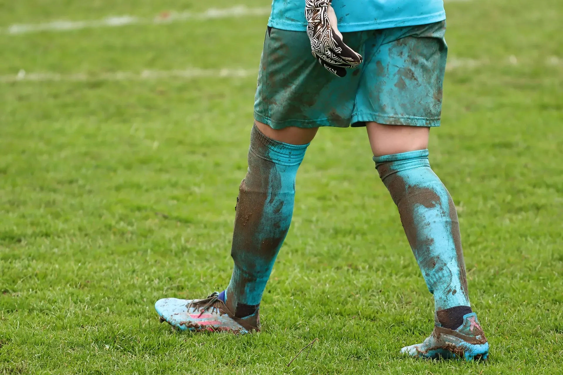 A goalkeeper in a mud-streaked teal kit walking across the pitch.